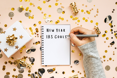 Hand writing "New Year IBS Goals" in a spiral notebook surrounded by gold confetti, ribbon, and a wrapped gift box.