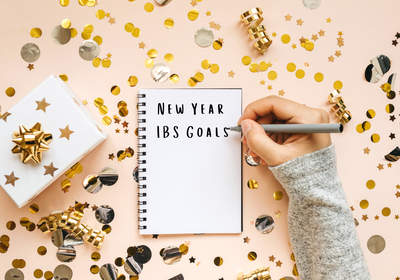 Hand writing "New Year IBS Goals" in a spiral notebook surrounded by gold confetti, ribbon, and a wrapped gift box.